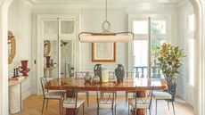 An elegant, sunlit dining room featuring a polished wood table, slender black metal chairs, and a unique horizontal pendant light. The room is decorated with ceramic vases and a large framed sketch on a paneled wall.