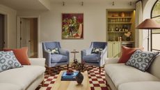 Cream living room with powder blue accent chairs and check brown and cream rug. There are large cream sofas either side, built in moss green alcove cabinets and shelving, and a large abstract green and orange framed art work centrally on the back wall