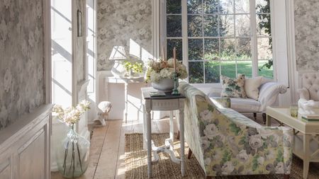 A floral living room with floral wallpaper, floral couch, white coffee table, rattan rug, wooden flooring, large glass vase with white flowers, and large windows out onto a leafy garden.