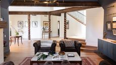Living room space with wooden pillars and beams, two leather armchairs, a lined coffee table with an open magazine, blue walls and cabinets. Pictured behind is a large open space with a round side table and framed pictures.