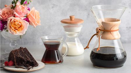 A Chemex Coffeemaker filled with black coffee, with a glass mug filled with coffee, glass milk holder, and a small plate of chocolate cake. Behind is a glass vase of roses, and the background is grey.